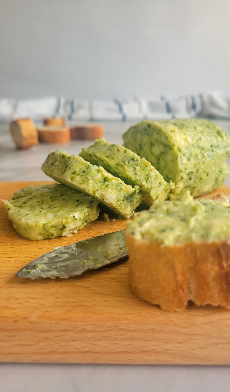 log of pesto butter on a cutting board, some cut into slices, bread in the front and back
