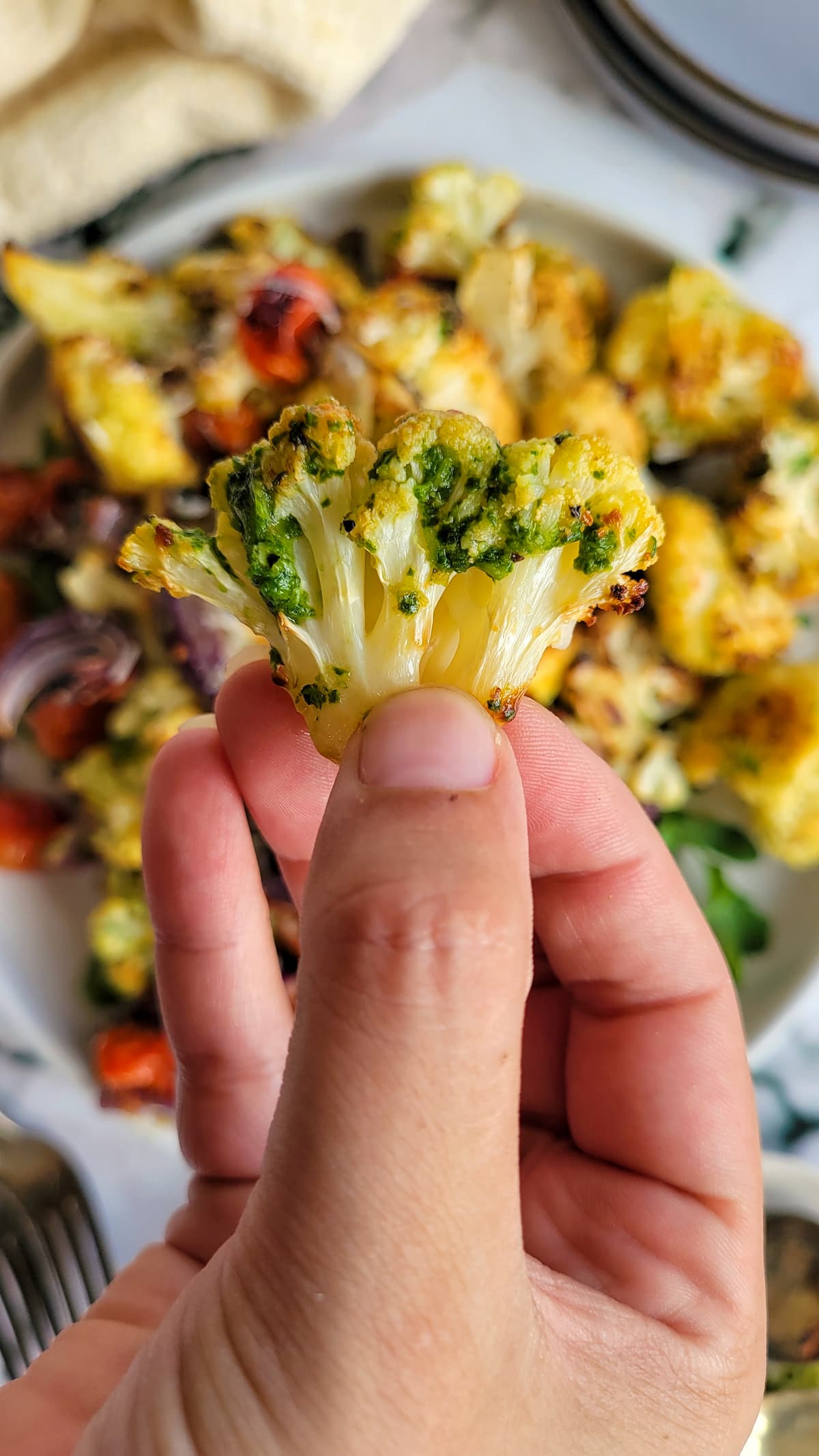 hand holding a piece of cooked cauliflower covered in pesto over a plate with more