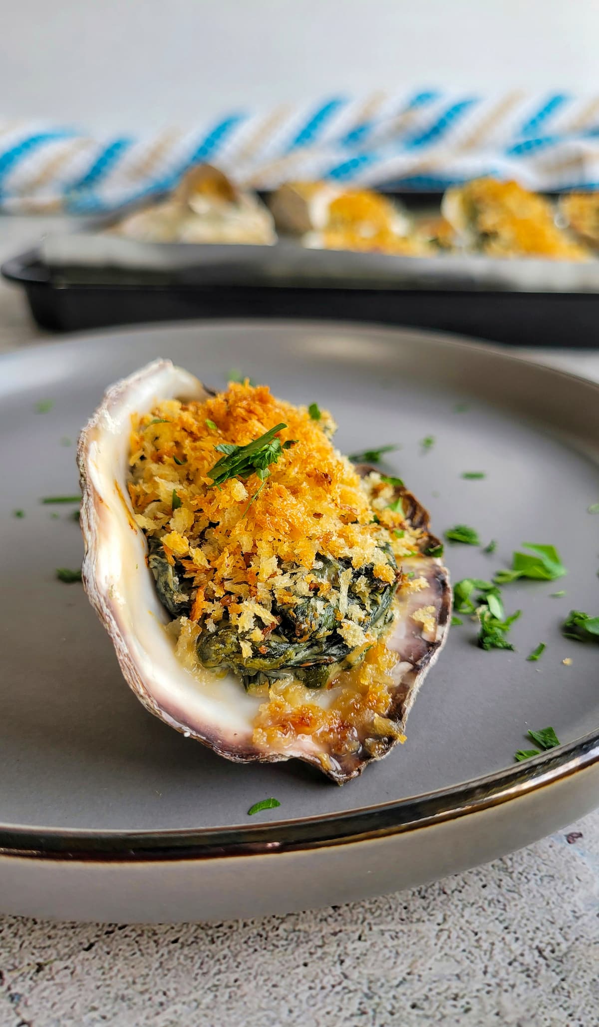 1 baked oyster with parsley, spinach, and breadcrumbs on a plate, more on a tray in the background
