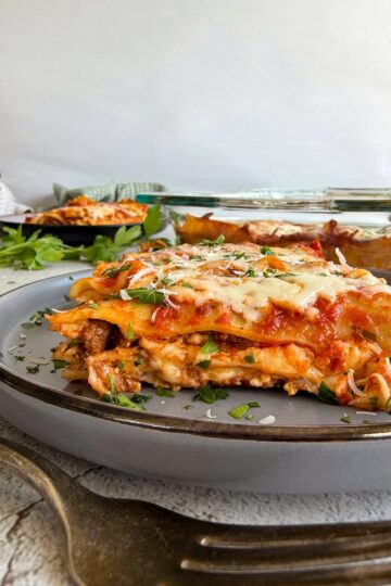 slice of lasagna with beef and ricotta on a plate garnished with fresh parm and parsley, another slice on a plate in the background