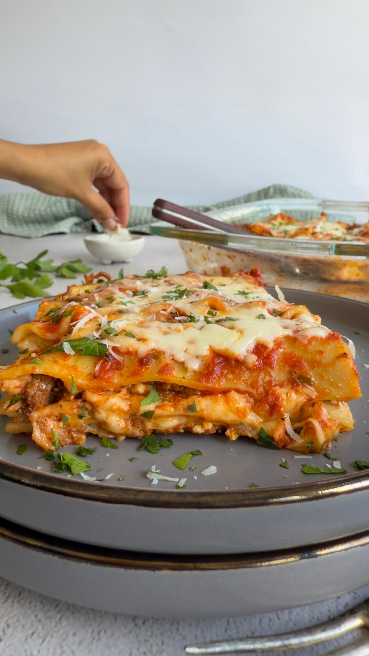 slice of lasagna on a plate garnished with fresh parm and parsley, hand grabbing some parm out of a small dish in the background next to the dish of lasagna