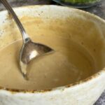 tahini caesar salad dressing in a bowl with a spoon, chopped romaine lettuce in a bowl behind it