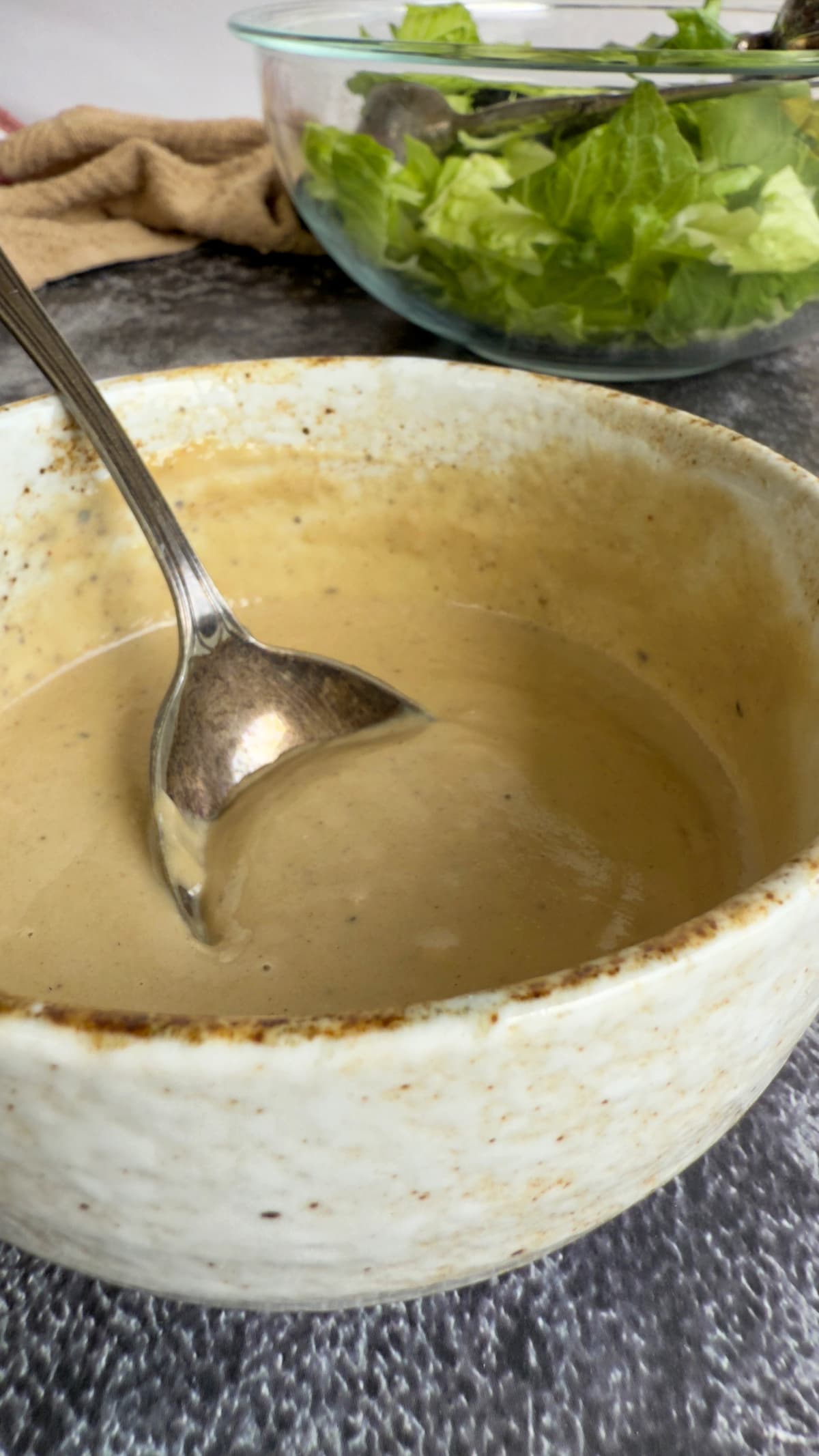 tahini caesar salad dressing in a bowl with a spoon, chopped romaine lettuce in a bowl behind it