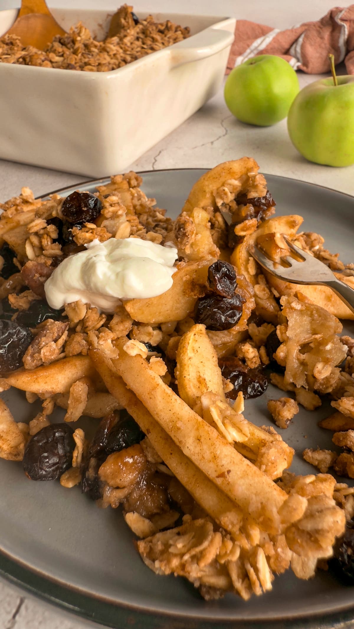 apple cranberry crisp on a plate with greek yogurt, oats and dried cranberries, dish with more in the background next to 2 apples