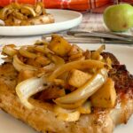 apples and onions on top of a cooked pork chop, another in the background next to an apple