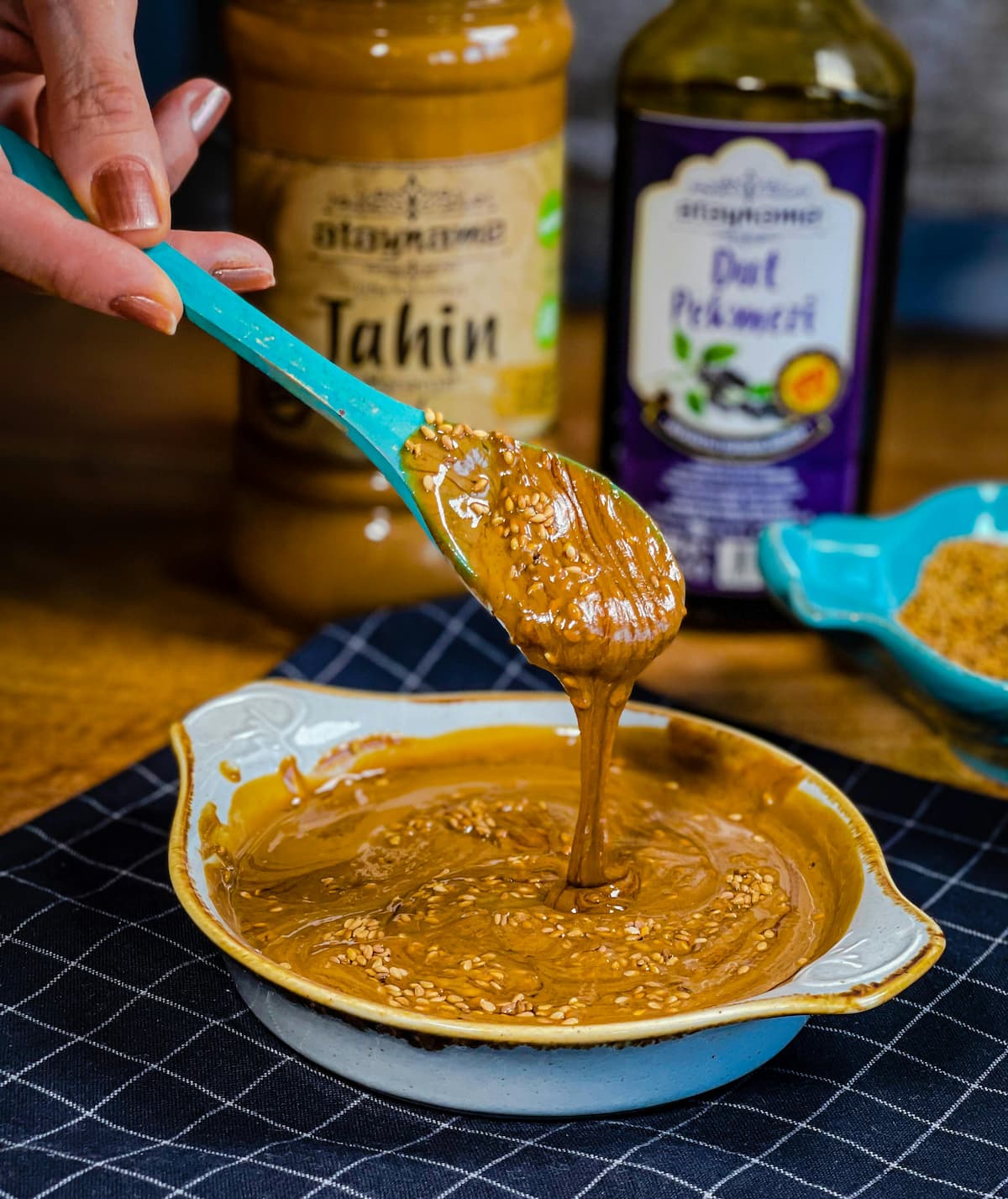 hand lifting some sesame paste out of a bowl with a spoon, jar of tahin and something else in the background
