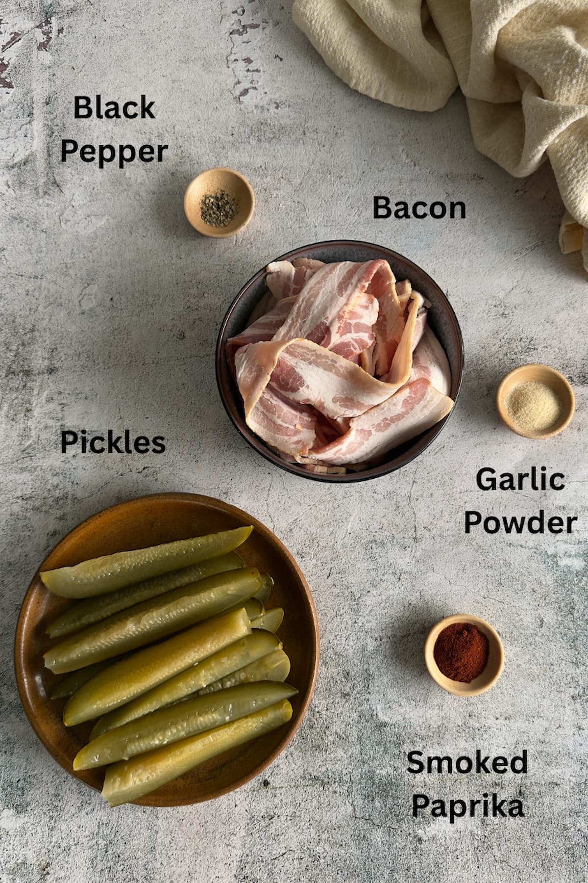ingredients for bacon wrapped dill pickles laid out in separate bowls - raw thin sliced bacon, dill pickle spears, garlic powder, smoked paprika, and black pepper.