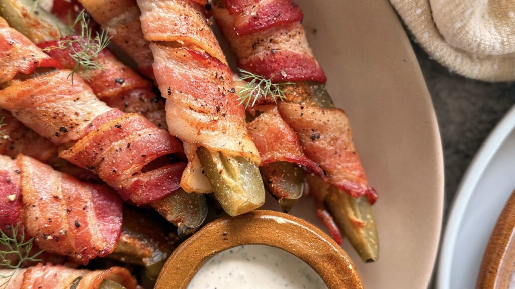 bacon wrapped dill pickles on a plate garnished with fresh cracked pepper and fresh dill. ramekin of white ranch dipping sauce also on the plate.