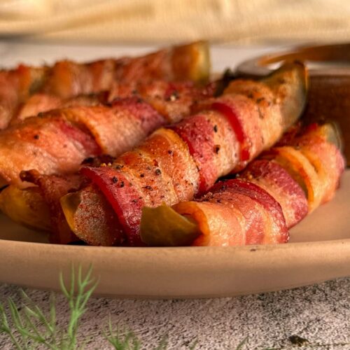 bacon wrapped pickle spears stacked on a plate. fresh dill in the front.