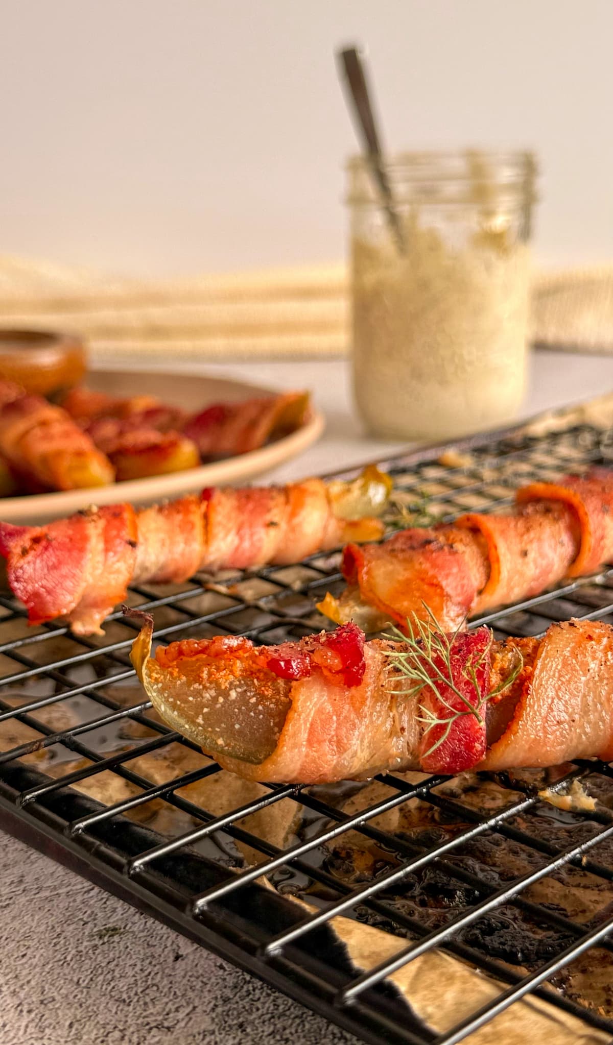bacon wrapped pickles on a wire rack over a parchment lined baking sheet, more on a plate in the background. Jar of ranch sauce with a spoon in it behind the tray.