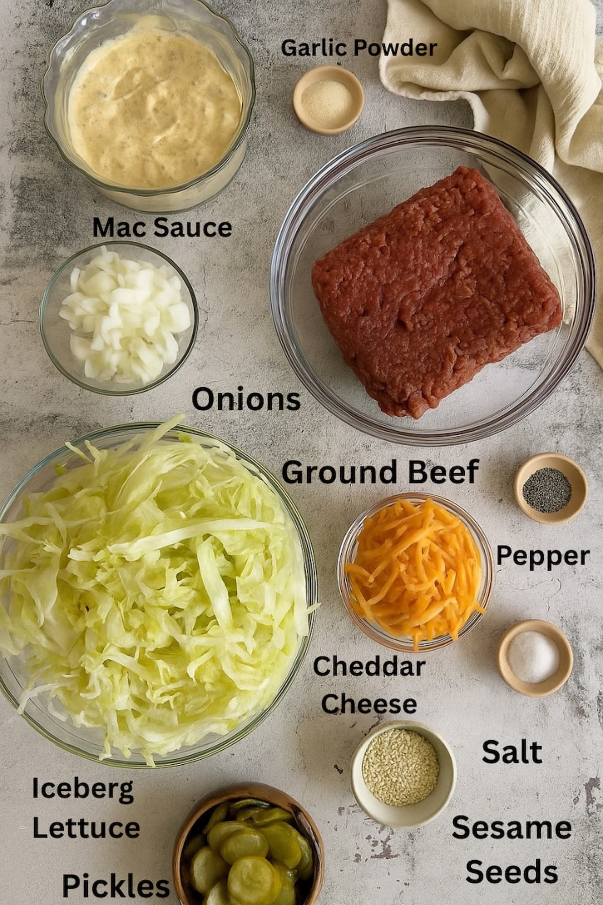 ingredients for big mac salad laid out in separate bowls - mac sauce, onions, ground beef, cheddar cheese, iceberg lettuce, garlic powder, salt, pepper, sesame seeds, pickles.