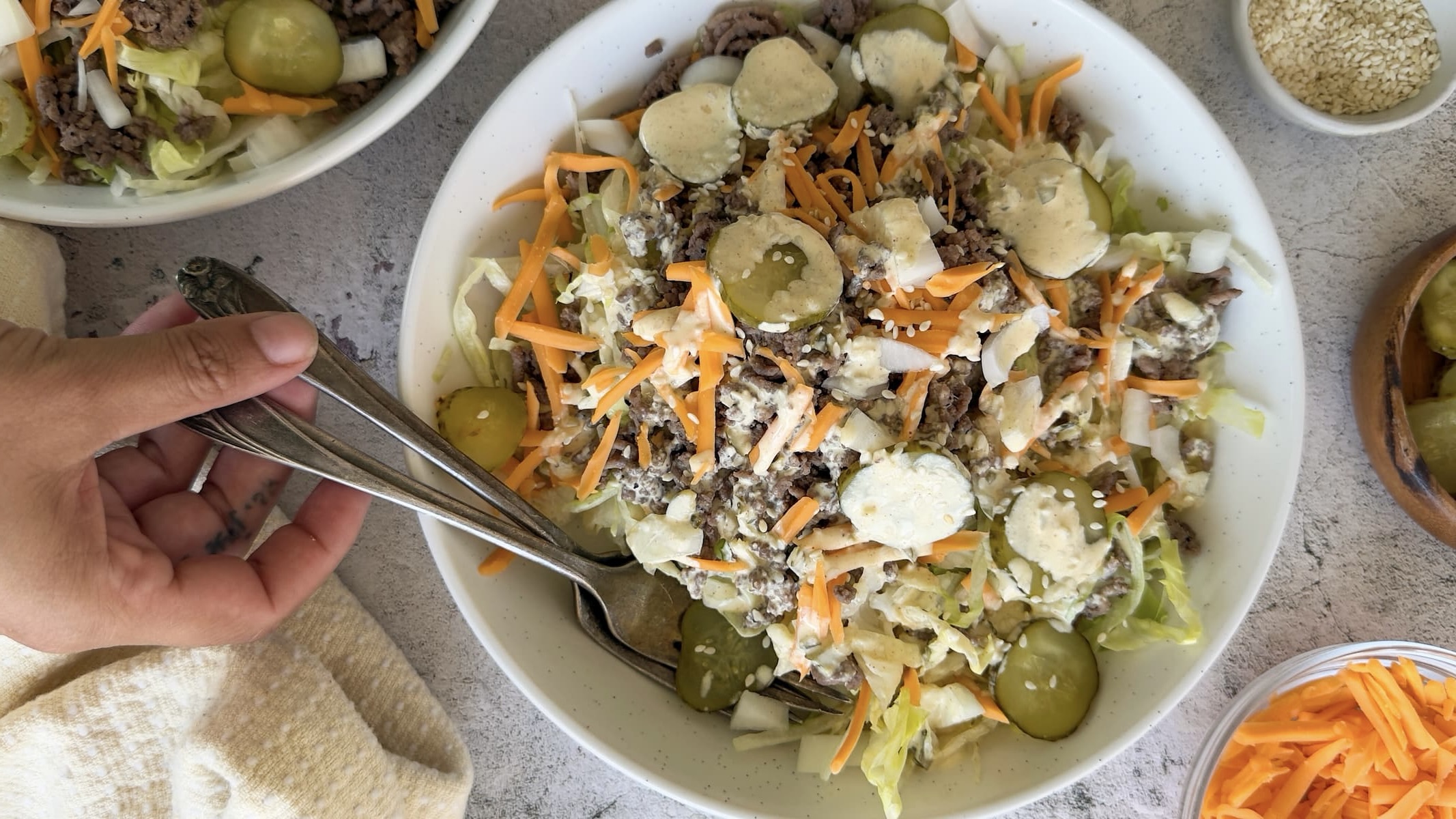 hand holding two forks in a bowl of a big mac salad with sliced pickles, grated cheddar, chopped lettuce, ground beef, sesame seeds, and a creamy mac sauce on top. Another salad and small bowls of toppings in the background.