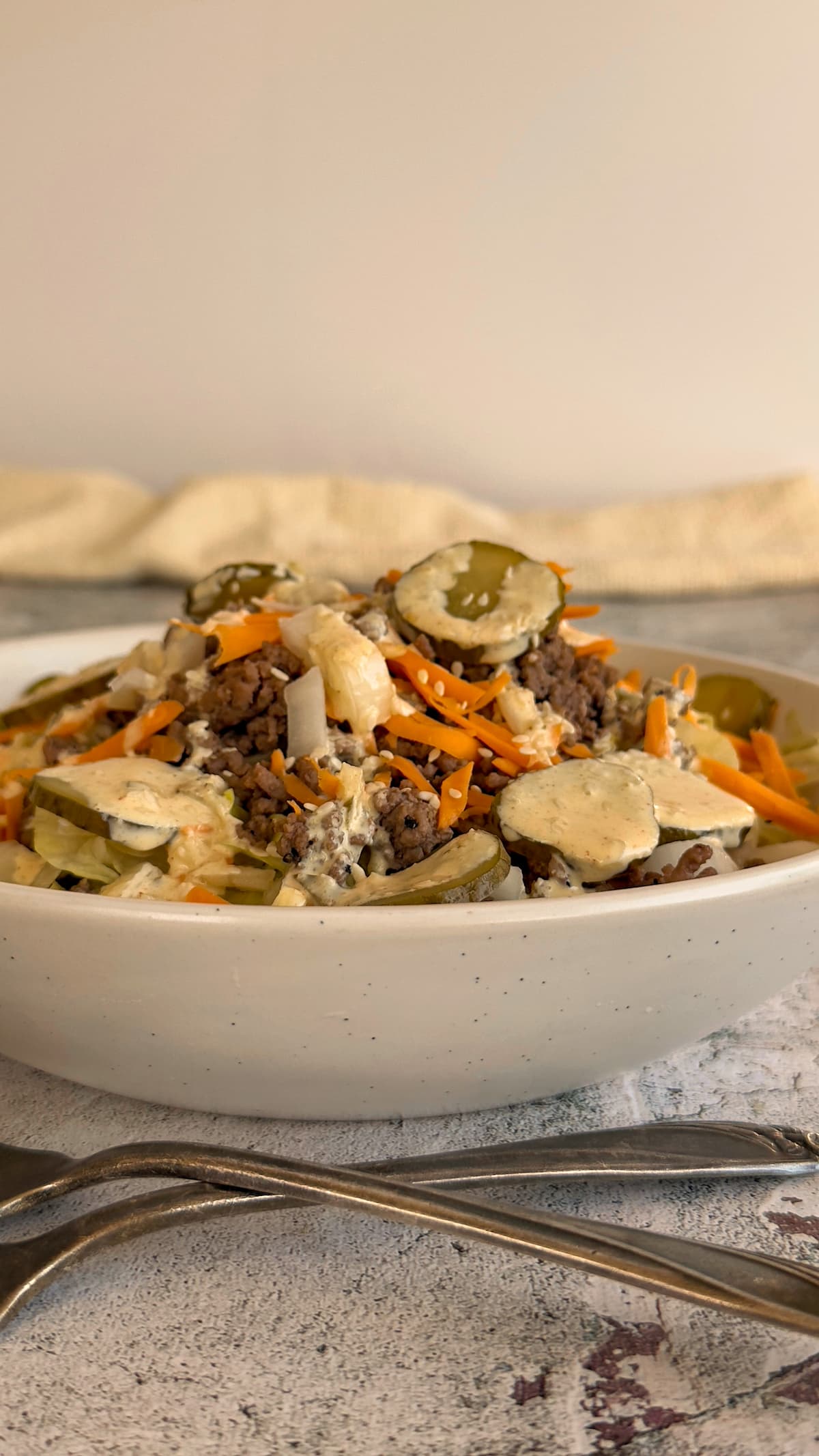 side view of a bowl with a big mac salad with cheddar cheese, ground beef, sesame seeds, and pickles piled high, topped with a creamy orange dressing.