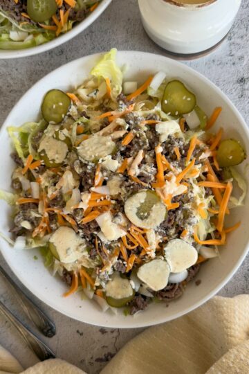 big mac salad in a bowl with shredded lettuce, sliced pickles, grated cheddar, sesame seeds, and a creamy mac sauce drizzled on top. Another salad and more sauce in a pourable container behind it.
