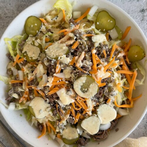 big mac salad in a bowl with shredded lettuce, sliced pickles, grated cheddar, sesame seeds, and a creamy mac sauce drizzled on top. Another salad and more sauce in a pourable container behind it.