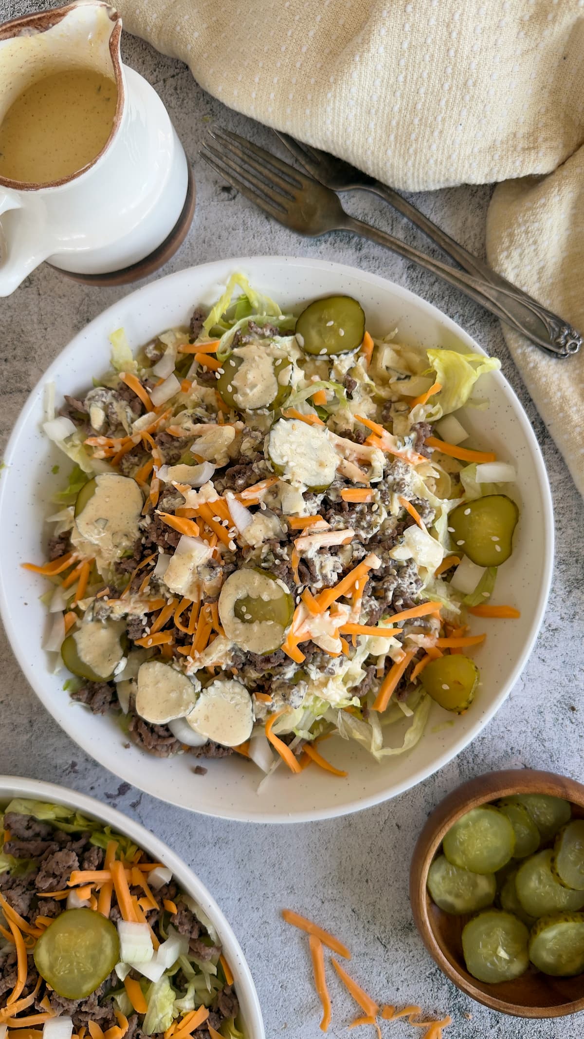 big mac salad in a bowl drizzled with mac sauce and sesame seeds. Sauce in a pourable container, another salad, and a bowl of sliced pickles in the background.