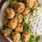 greek chicken meatballs sprinkled with lemon zest stacked on a bed of arugula on a plate. Blob of tzatziki sauce on the plate, plus more in the background with a spoon on a separate plate.