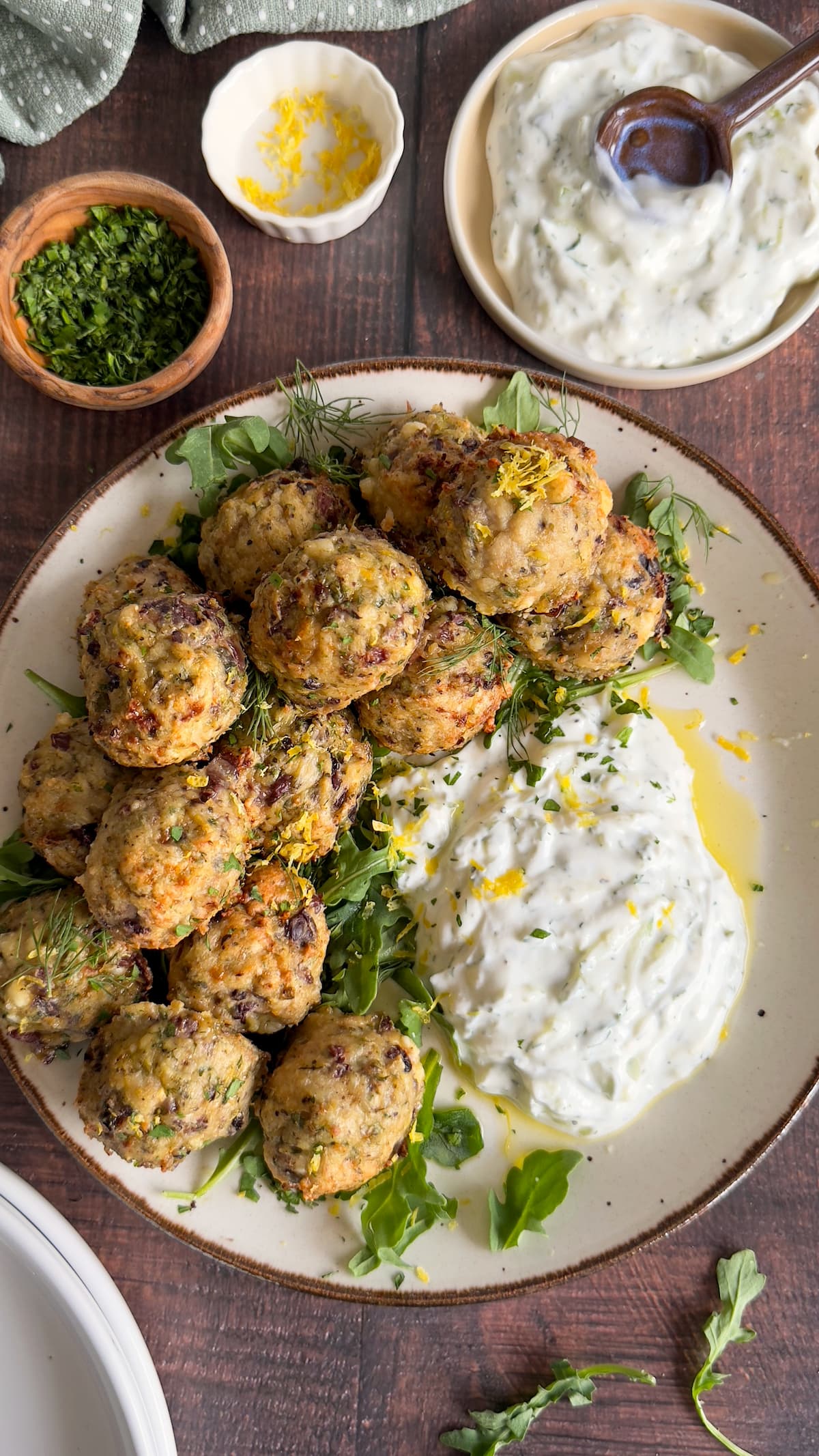 plate of greek meatballs with arugula, lemon zest, tzatziki sauce. More lemon zest, herbs, and tzatziki in separate bowls around.