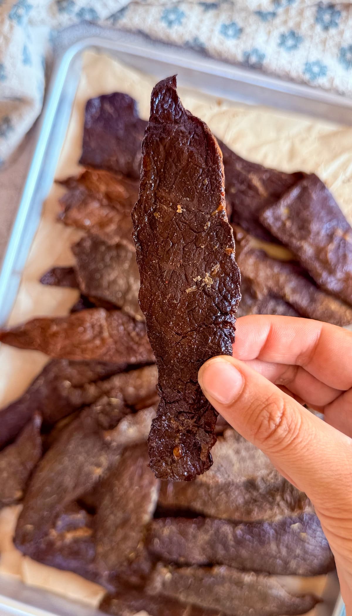 hand holding a piece of beef jerky over a sheet pan with more.