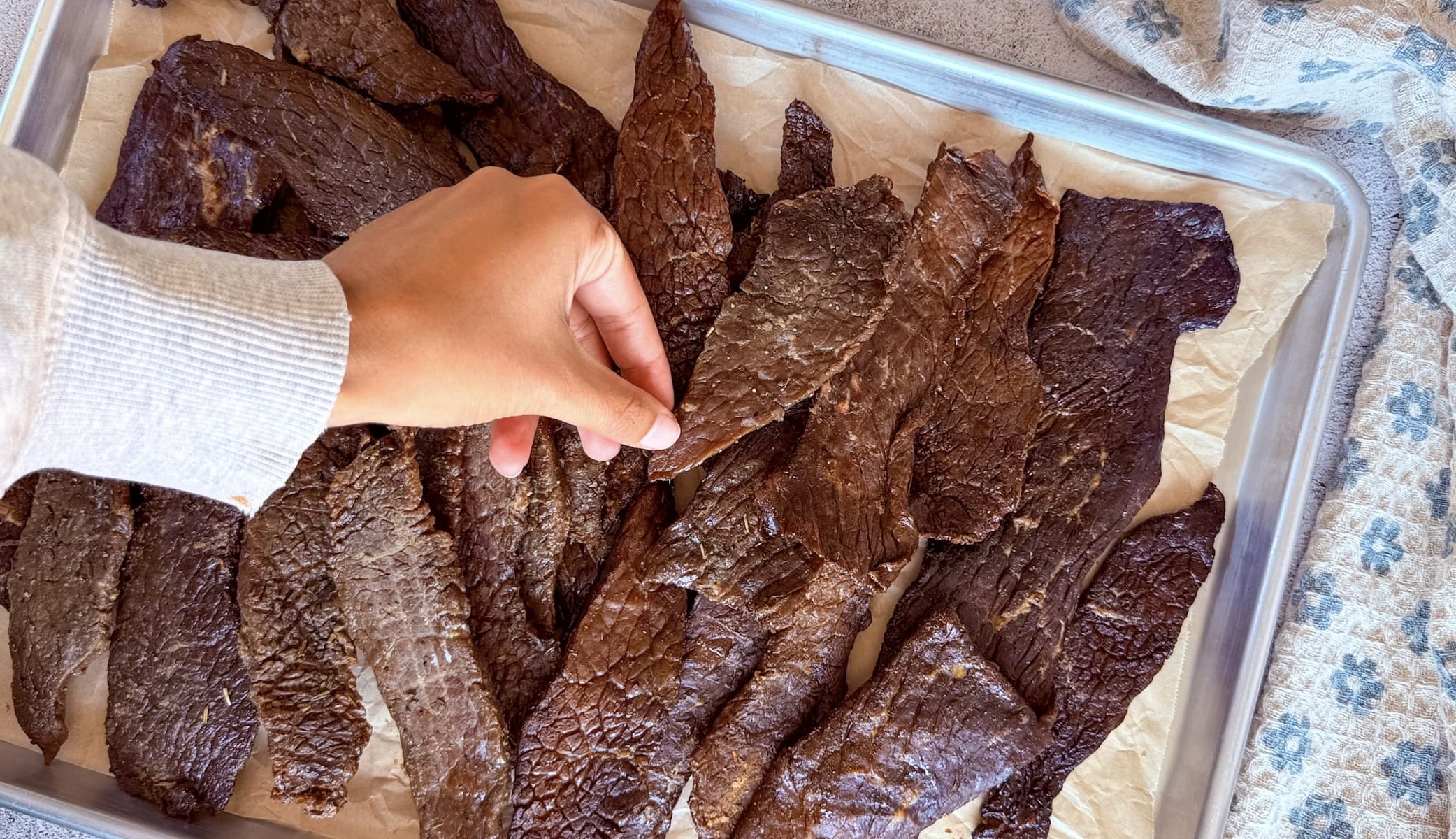 hand grabbing a piece of beef jerky off a parchment lined baking sheet with a lot more.