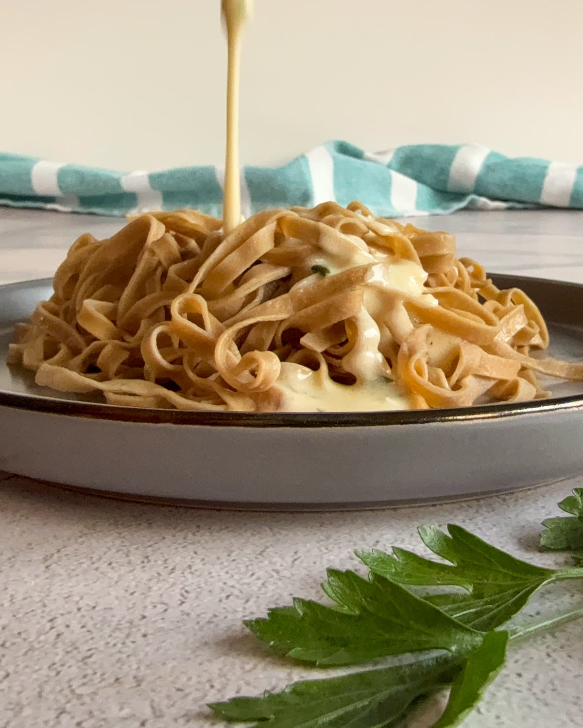 creamy alfredo sauce pouring onto a plate of fettuccine, sprig of fresh parsley in front of the plate.