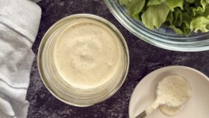 peppercorn ranch dressing in a jar next to a white plate with a spoon full of the dressing on it. A bowl of chopped lettuce in the background.