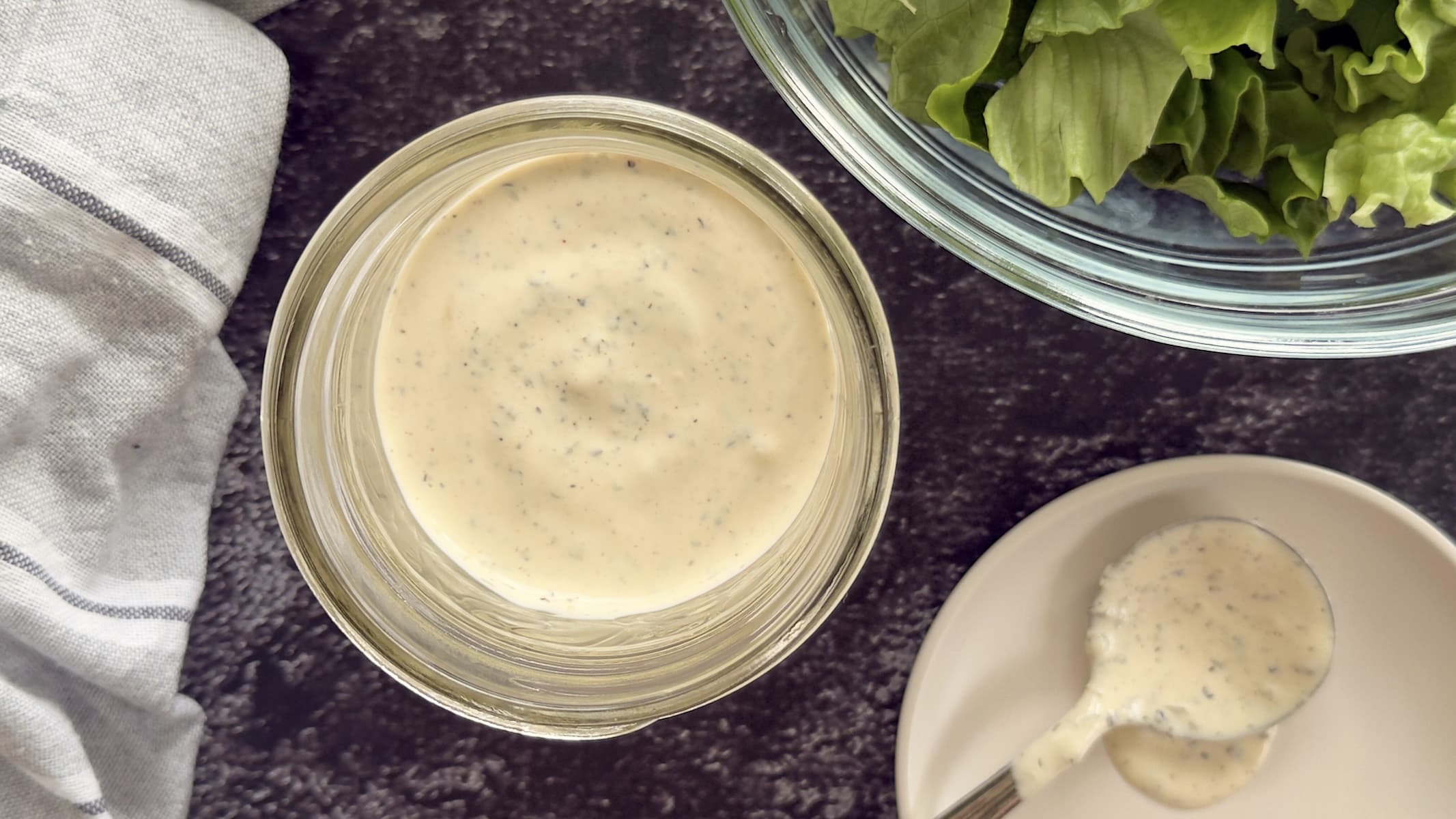 peppercorn ranch dressing in a jar next to a white plate with a spoon full of the dressing on it. A bowl of chopped lettuce in the background.