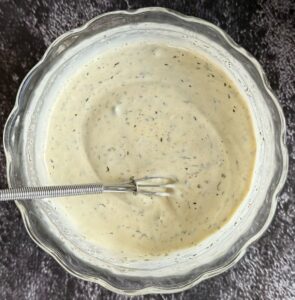 creamy white sauce in a bowl with specks of black peppercorn and other spices. Little whisk in the bowl.