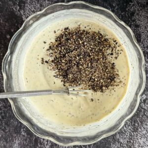 pile of freshly cracked black peppercorns on top of a creamy dressing in a bowl with a little whisk.