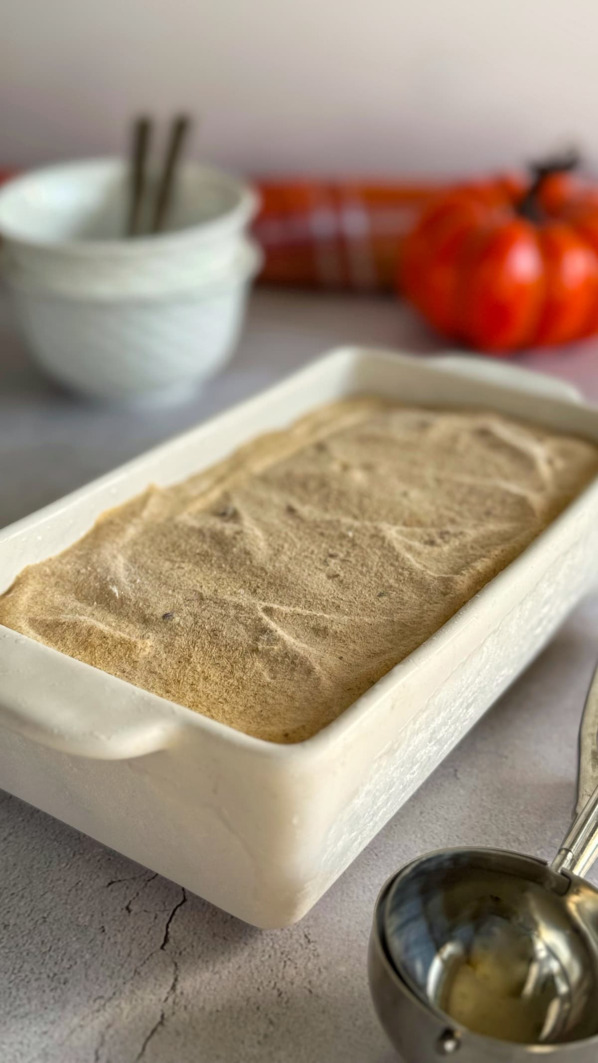 rectangular loaf pan with untouched pumpkin spice ice cream next to an ice cream scoop. bowls with spoons next to a pumpkin in the background.