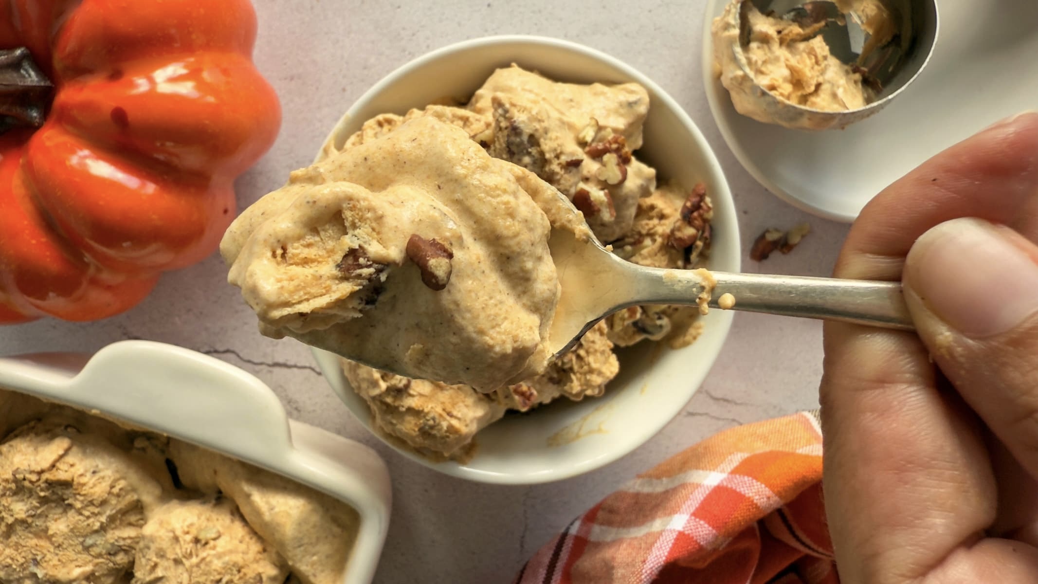 spoonful of pumpkin spice ice cream with chopped pecans over a bowl with more. pumpkin and container of more incream in the background.