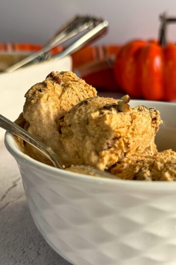 two scoops of pumpkin spice ice cream with pecans in a white bowl with a spoon, pumpkin in the background.