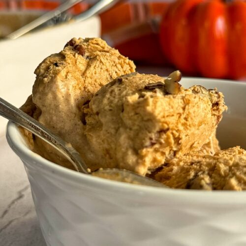 two scoops of pumpkin spice ice cream with pecans in a white bowl with a spoon, pumpkin in the background.