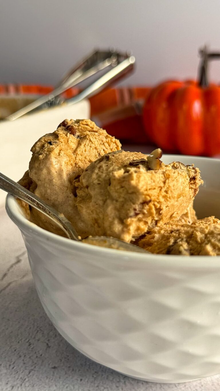 two scoops of pumpkin spice ice cream with pecans in a white bowl with a spoon, pumpkin in the background.