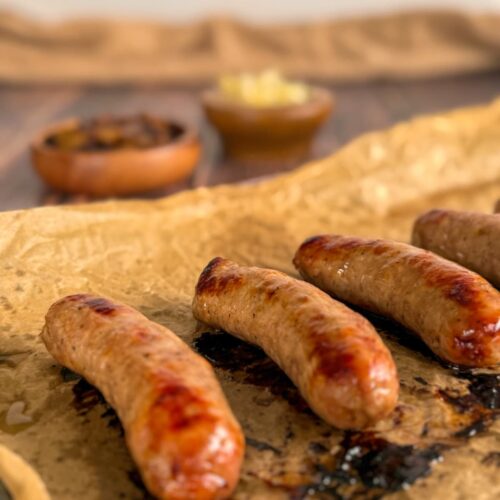4 cooked bratwurst on a parchment lined baking sheet. bowls of sauerkraut and caramelized onions in the background.