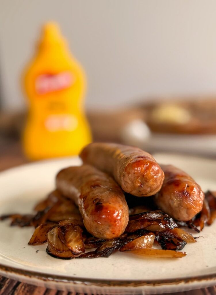 3 baked bratwurst stacked on top of a pile of caramelized onions on a plate. bottle of yellow mustard in the background.