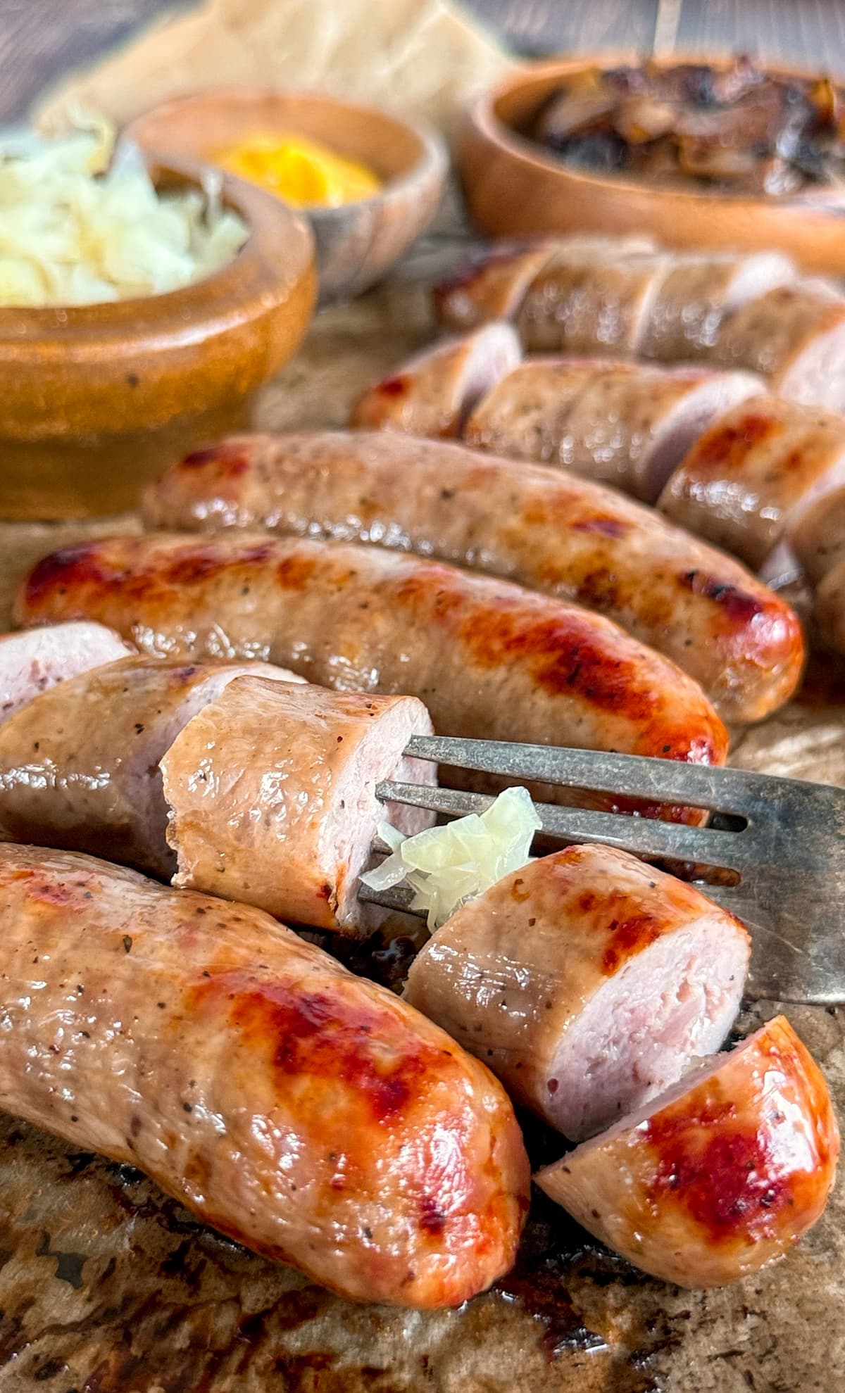 bratwurst sausage links on a parchment lined baking sheet - some sliced, some not. ramekins of caramelized onions, mustard, and sauerkraut on the tray. fork piercing a piece of sausage.