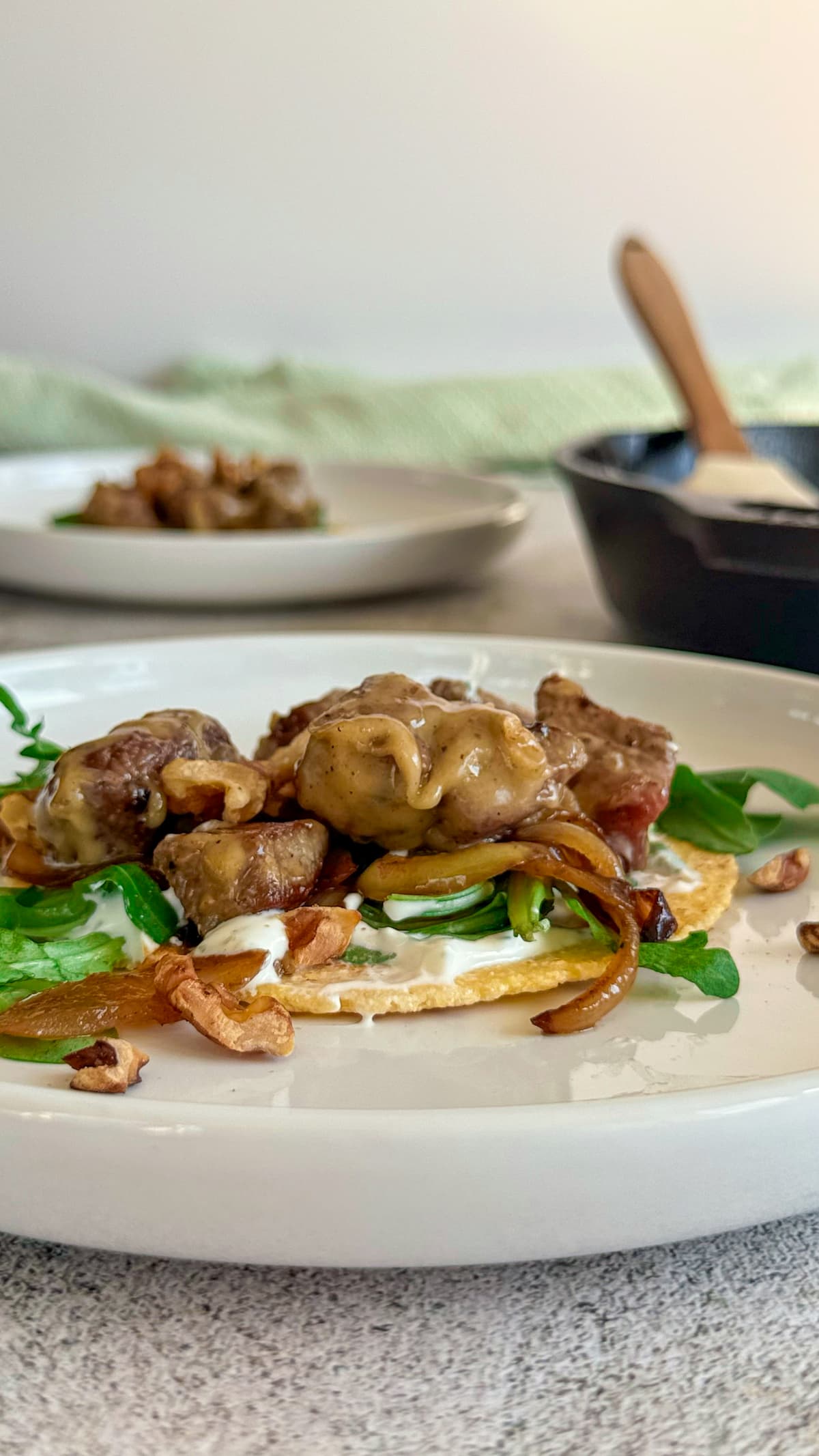 tortilla on a white plate topped with steak chunks, creamy brie cheese sauce, arugula, creamy white sauce, walnuts, and caramelized onions. another taco on a plate in the background next to a cast iron skillet with a rubber spatula in it.