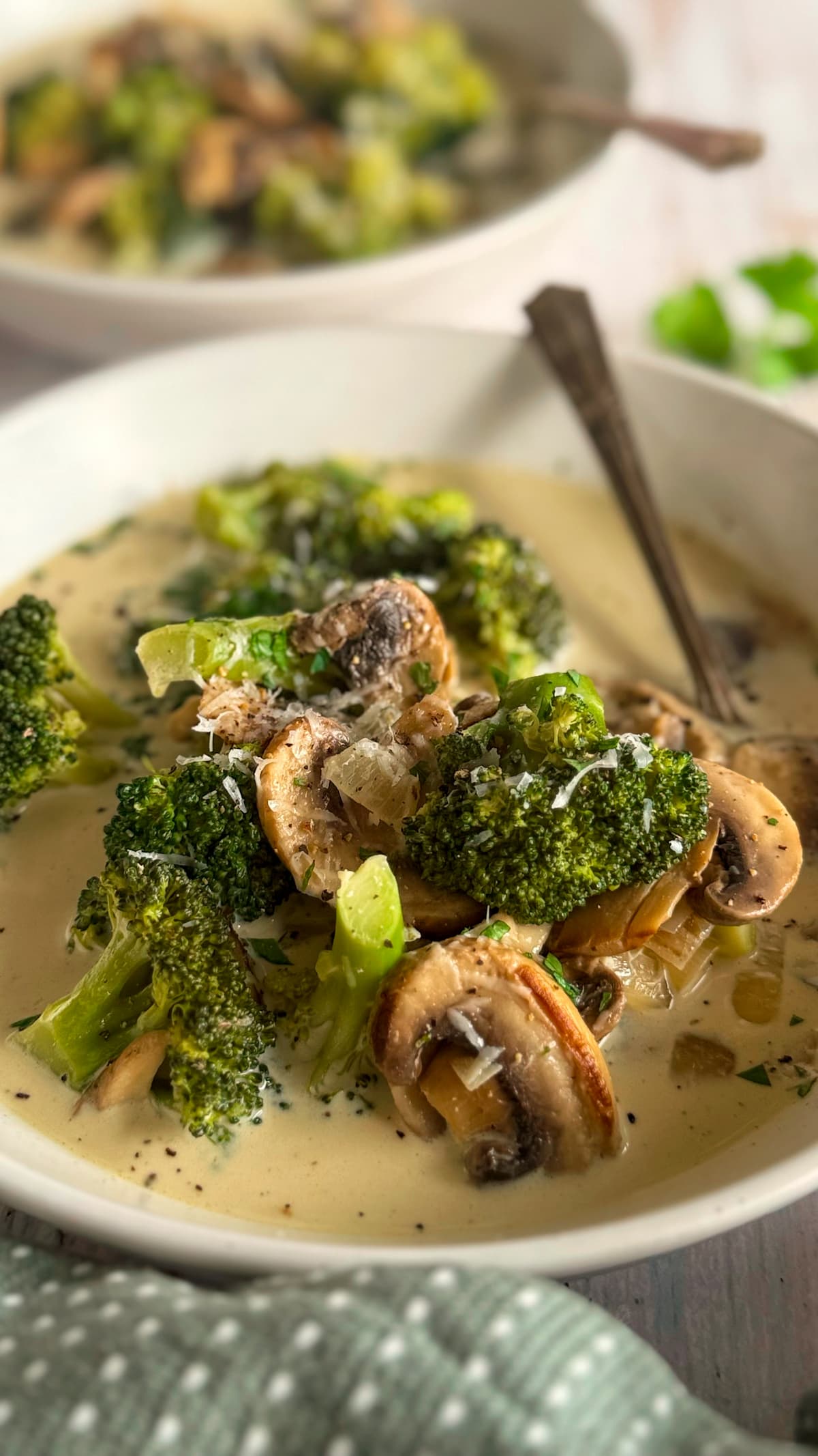 side view of a bowl of creamy mushroom broccoli soup, spoon in the bowl, another bowl of soup in the background.