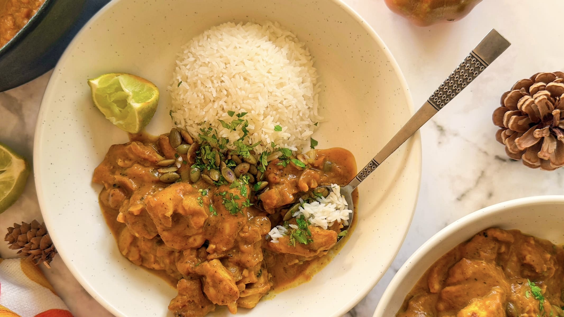 bowl of pumpkin and chicken curry topped with toasted pumpkin seeds and chopped cilantro, rice and lime wedge in the bowl with a spoon next to another bowl of curry and a pinecone.