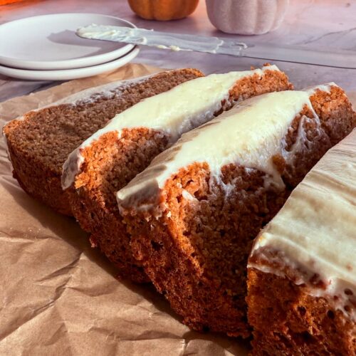 sliced frosted pumpkin bread on a piece of parchment paper. frosting knife on two small plates next to a couple coloured pumpkins in the background.