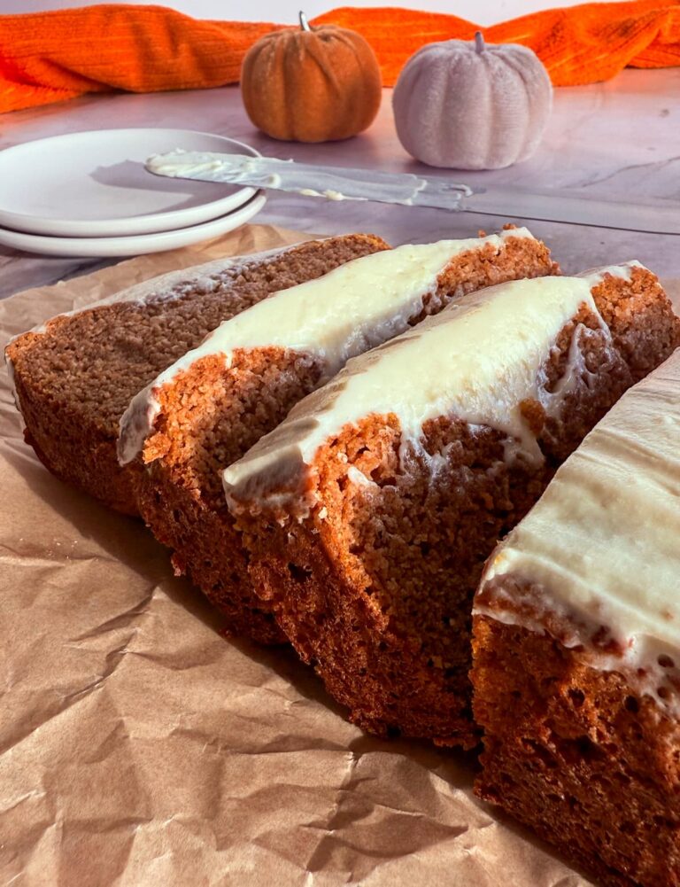 sliced frosted pumpkin bread on a piece of parchment paper. frosting knife on two small plates next to a couple coloured pumpkins in the background.