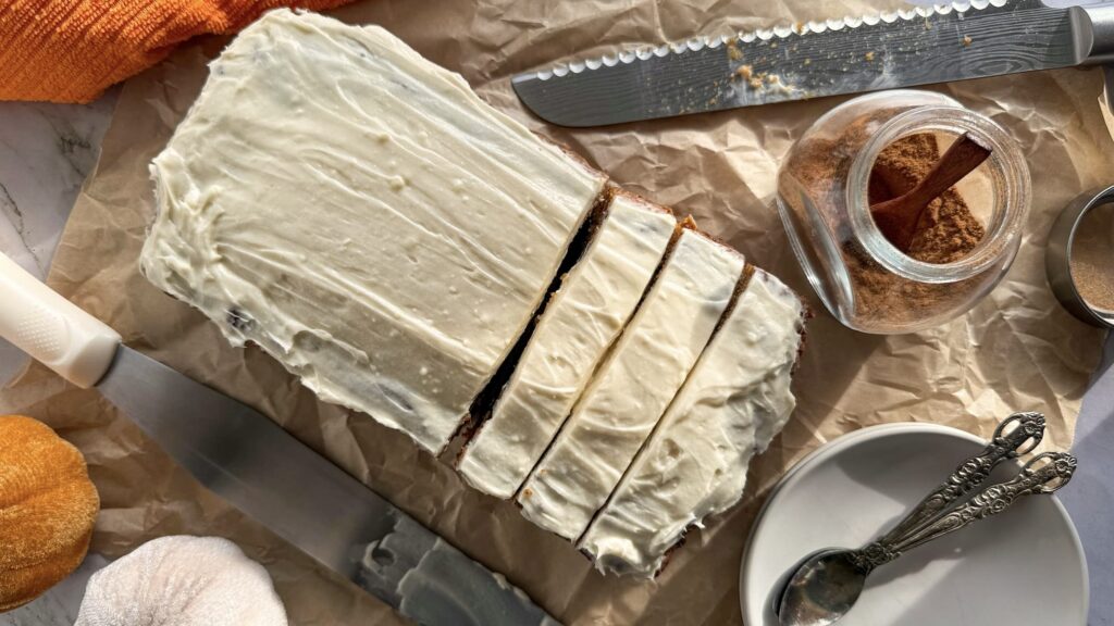 half sliced frosted pumpkin bread on parchment paper next to a jar of pumpkin spice, a plate with small spoons, a dirty bread knife and froster, and a couple of coloured pumpkins.