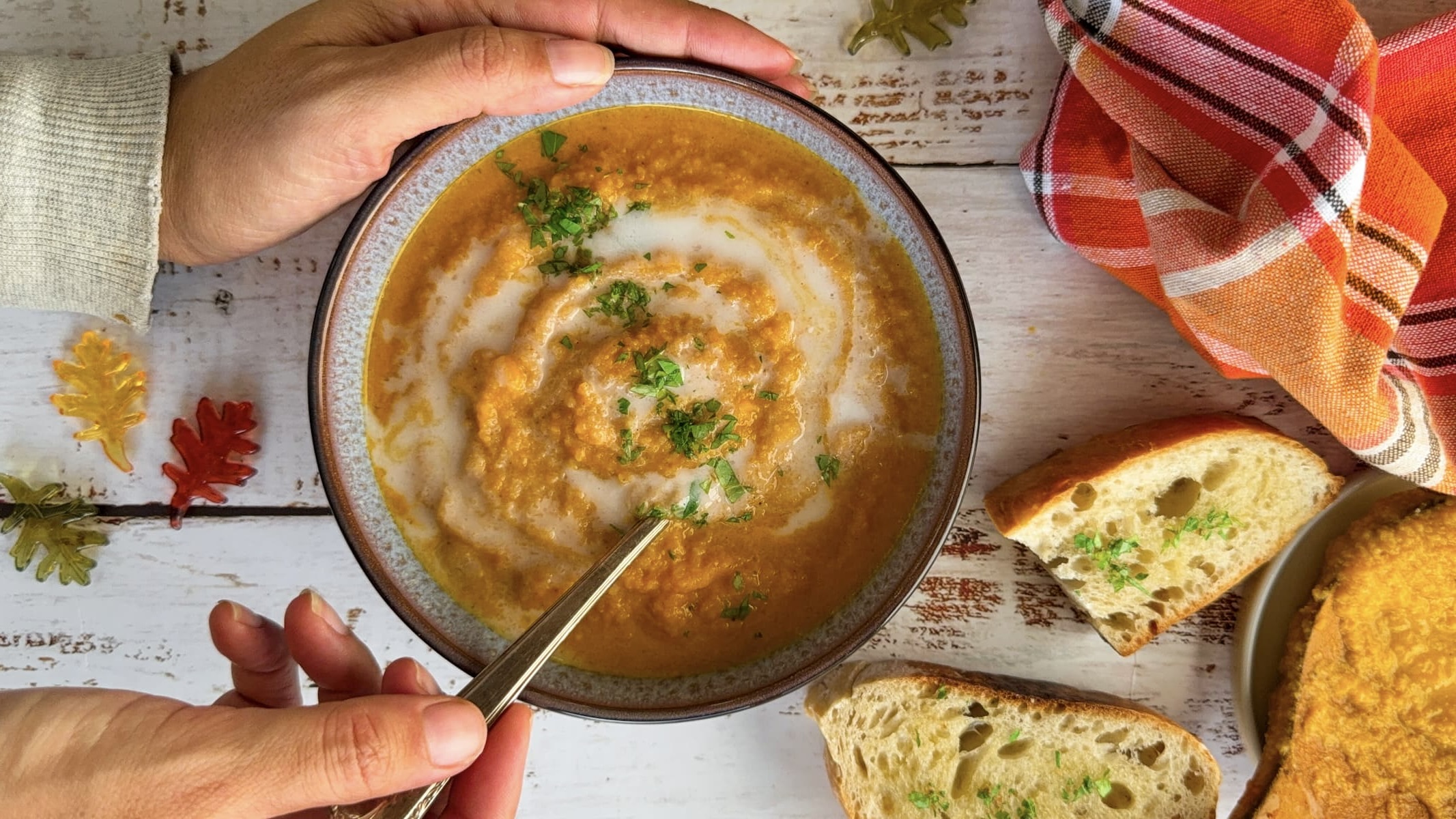 hands holding a bowl of cauliflower pumpkin soup swirled with coconut milk topped with chopped cilantro. two pieces of garlic bread next to it as well as fall colored leaves in the background.