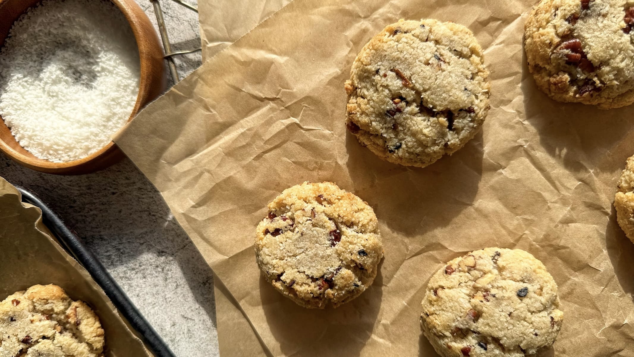 5 freshly baked coconut pecan cookies on a piece of parchment paper next to a bowl of shredded coconut. a parchment lined tray with more cookies beside.