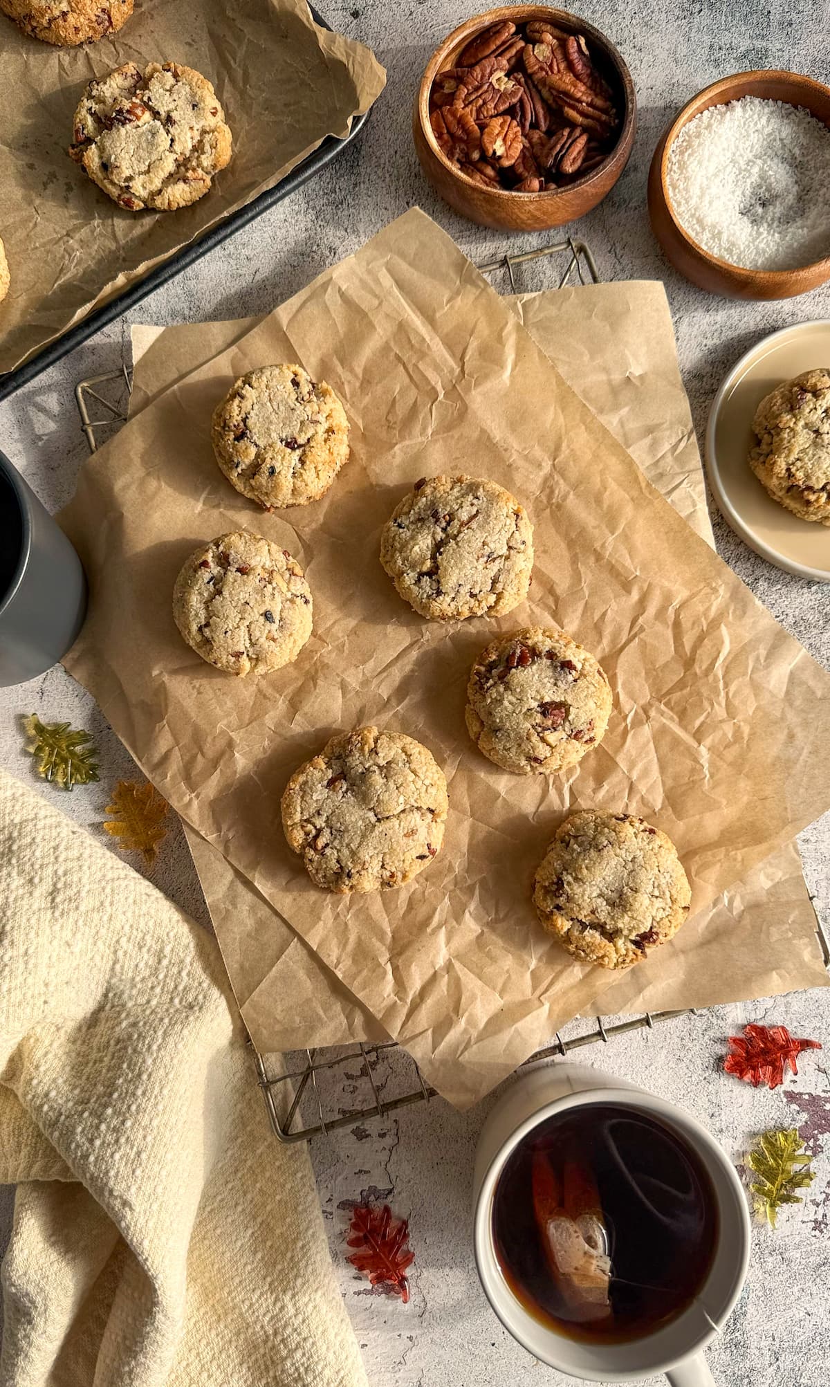 6 pecan cookies with coconut on a piece of parchment paper surrounded by bowls filled with ingredients (pecans, shredded coconut). 2 cups of tea in the background along with a cookie on a plate and more cookies on a parchment lined tray.