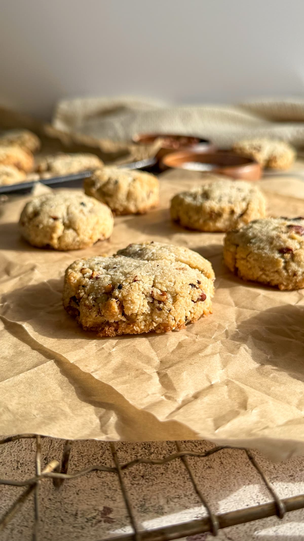 side view of freshly baked coconut cookies on a double piece of parchment paper over a wire rack. more cookies on a tray in the background next to bowls of ingredients.