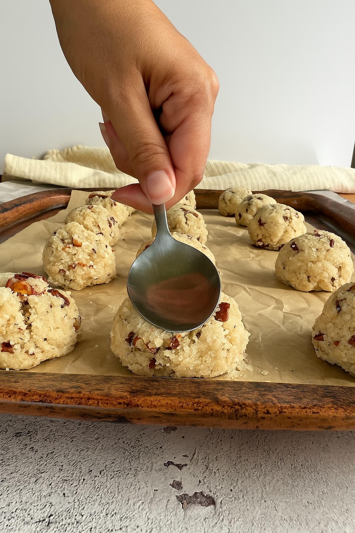 hand with a spoon pressing down on the top of an unbaked cookie to flatten it. more unbaked cookies on the parchment lined tray.