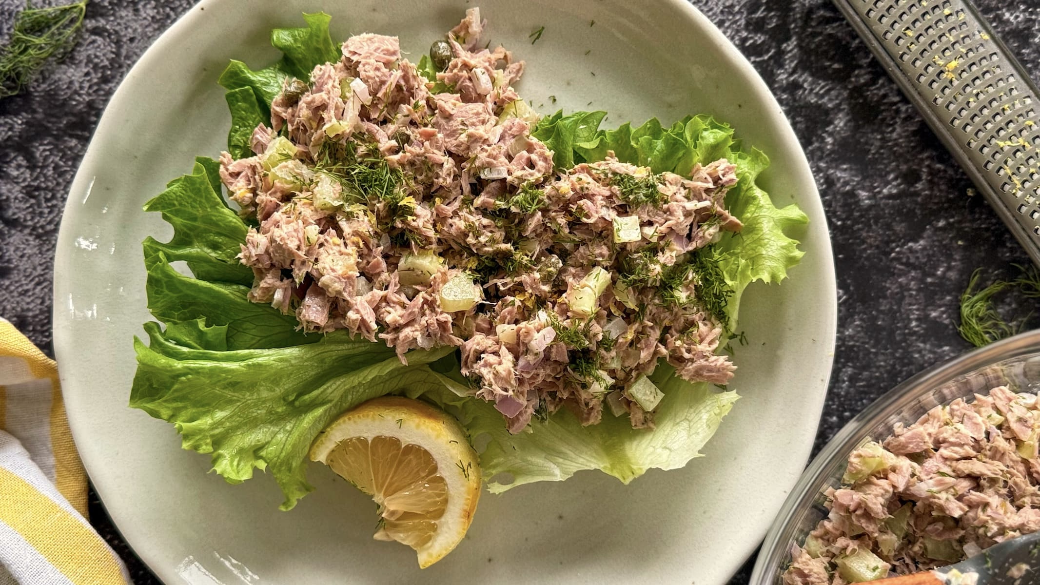 plate with lemon and dill tuna salad in leaf lettuce cups with a lemon wedge. more tuna salad in a bowl next to it with a lemon zested microplane grater.