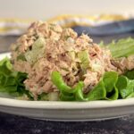 tuna salad with celery, shallots, and fresh dill stacked on leaf lettuce on a white plate. a few pieces of fresh dill in the front.